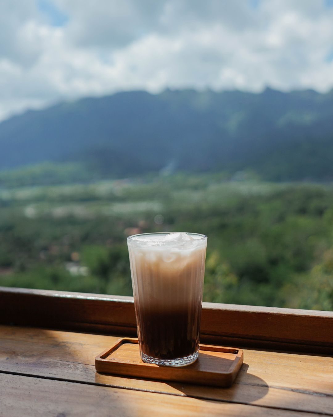 Singkong Keju & Kopi di Kedai Bukit Rhema: Cita Rasa Perbukitan Menoreh 1 Es kopi susu di pagar kayu Kedai Bukit Rhema dengan latar Perbukitan Menoreh